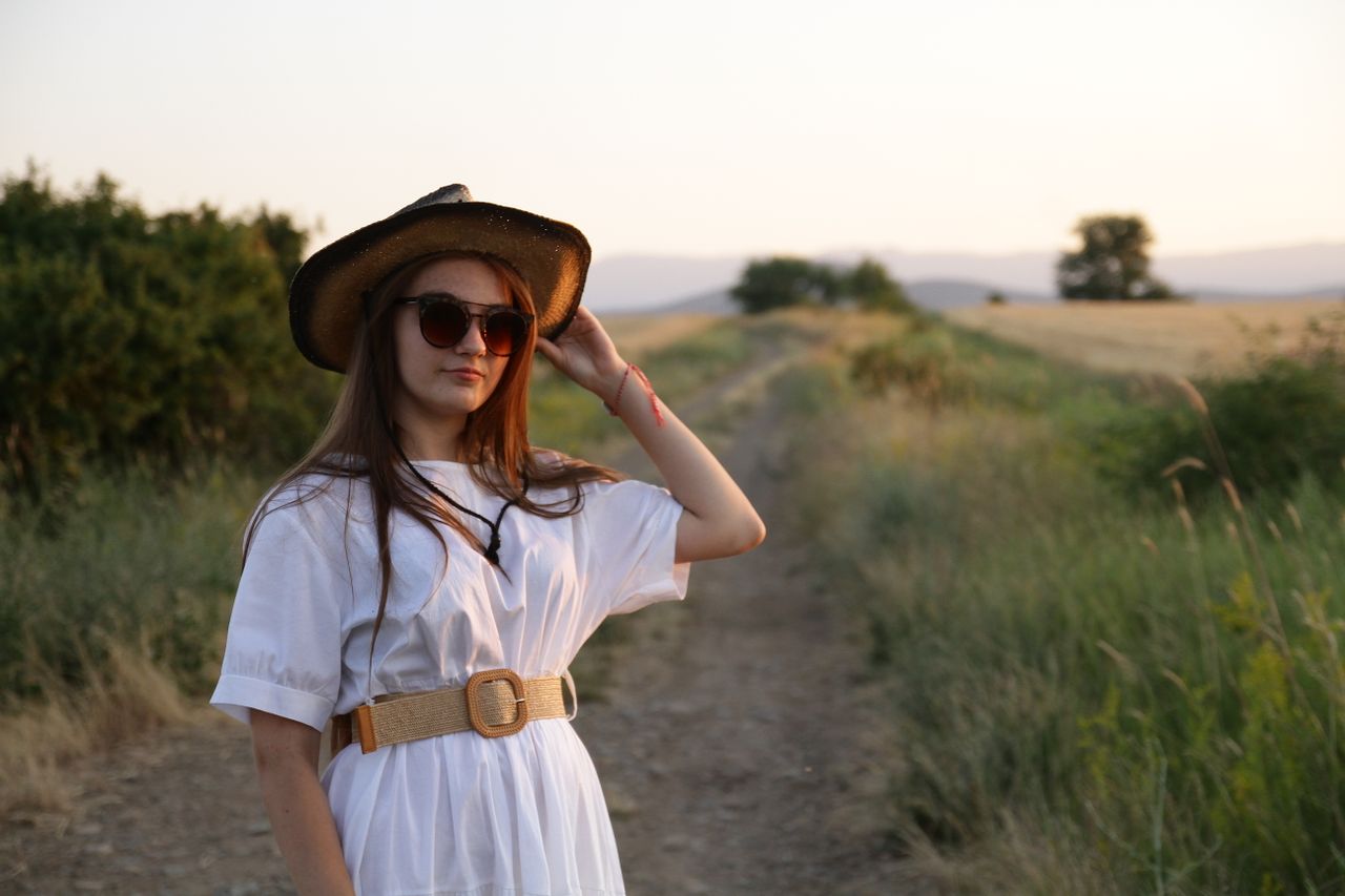 our-story A young woman in sunglasses and a straw hat poses on a rural path at sunset.