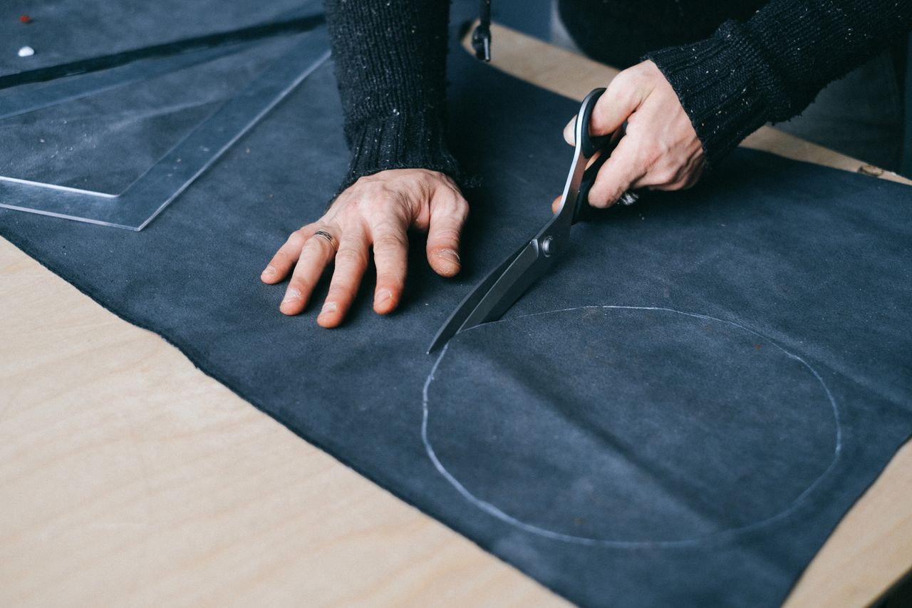 about-us High angle of crop anonymous male tailor with scissors cutting round piece of cloth while working in atelier